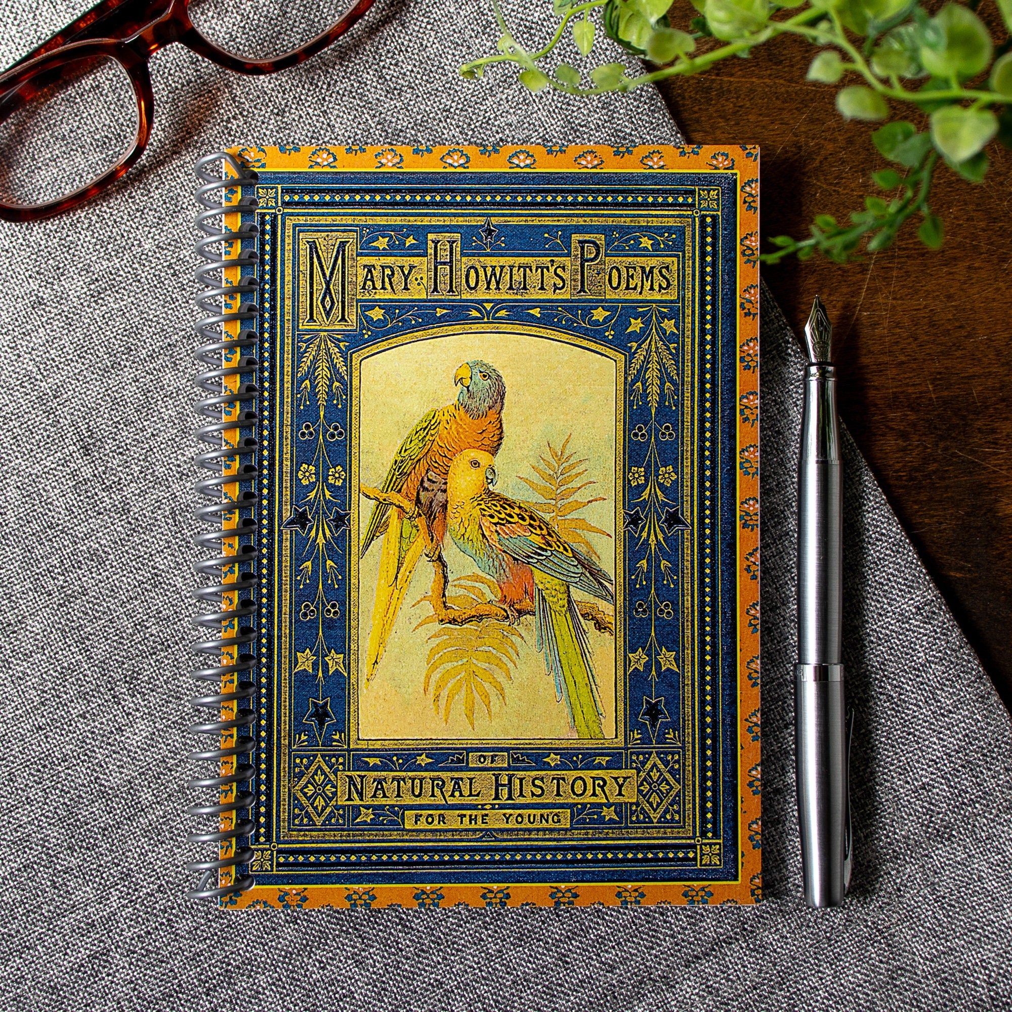 A travel-sized notebook featuring a vintage book cover: Mary Howitt&#39;s Poems of Natural History for the Young. Features a framed illustration of two colorful parrots. Colors are deep blue, orange, gold and green.