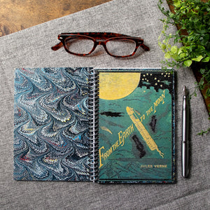 A travel-sized spiral notebook featuring a vintage book cover: From the Earth to the Moon by Jules Verne. Features an illustration of a rocket ship flying towards the moon. The colors are deep green, blue, black, and gold.