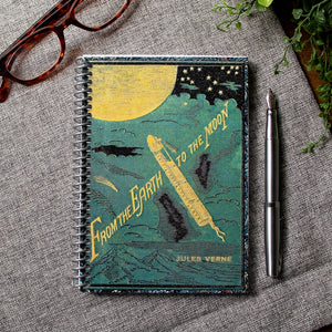 A travel-sized spiral notebook featuring a vintage book cover: From the Earth to the Moon by Jules Verne. Features an illustration of a rocket ship flying towards the moon. The colors are deep green, blue, black, and gold.