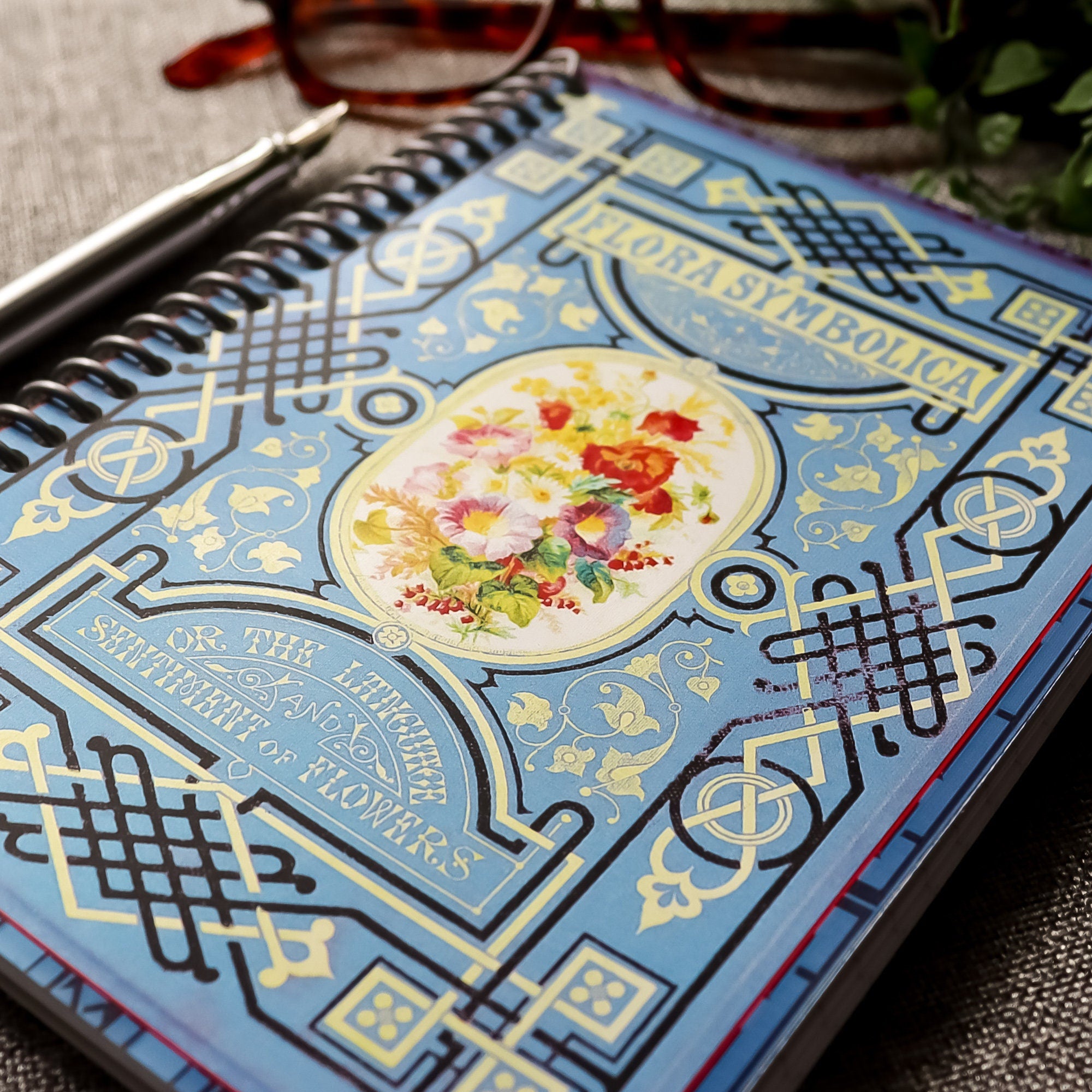 A travel-sized spiral notebook featuring a vintage book cover: Flora Symbolica or The Language and Sentiment of Flowers. Features a floral illustration surrounded by an ornate frame. The colors are sky blue, black, red, pink and gold.