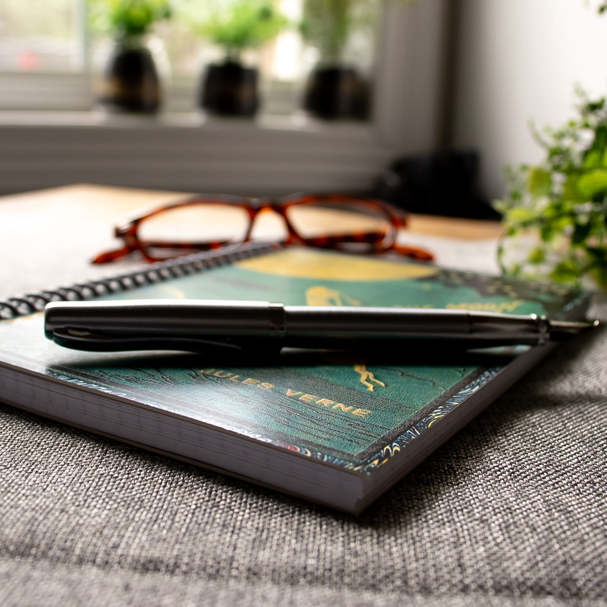 Close up image of a travel-sized spiral notebook featuring a vintage book cover: From the Earth to the Moon by Jules Verne. Features an illustration of a rocket ship flying towards the moon. The colors are deep green, blue, black, and gold.