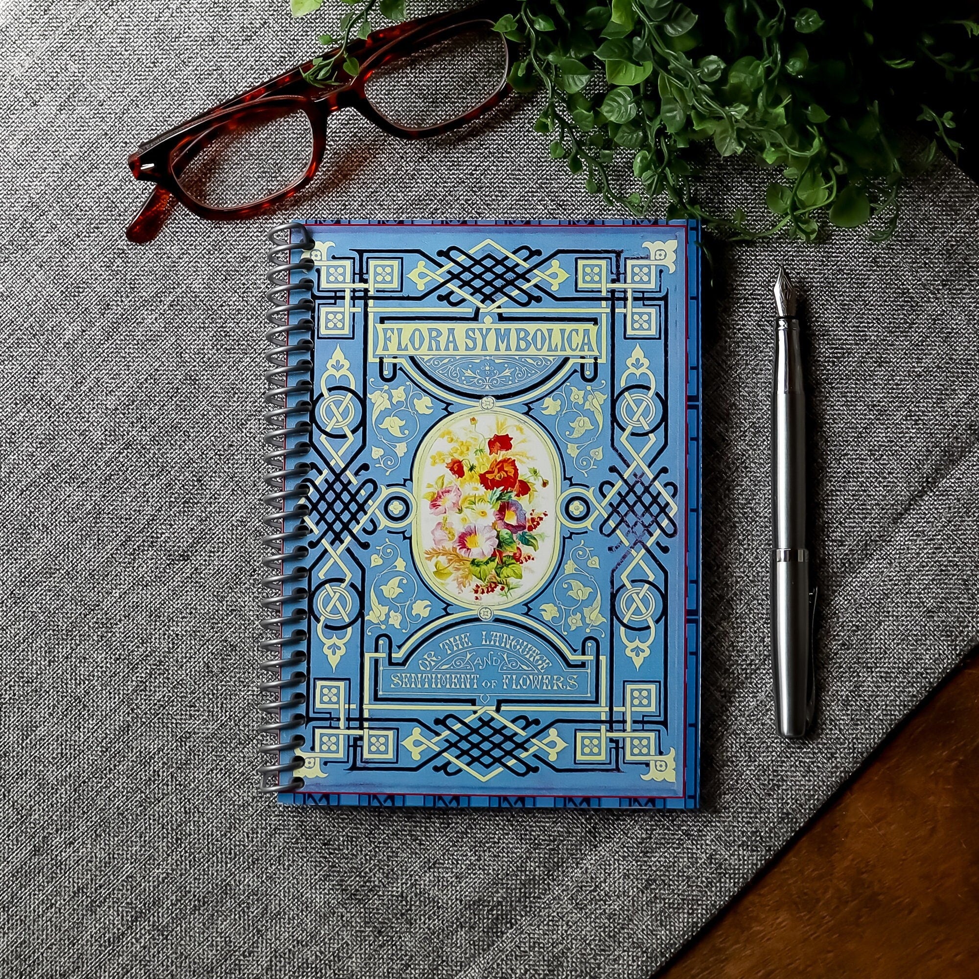 A travel-sized spiral notebook featuring a vintage book cover: Flora Symbolica or The Language and Sentiment of Flowers. Features a floral illustration surrounded by an ornate frame. The colors are sky blue, black, red, pink and gold.