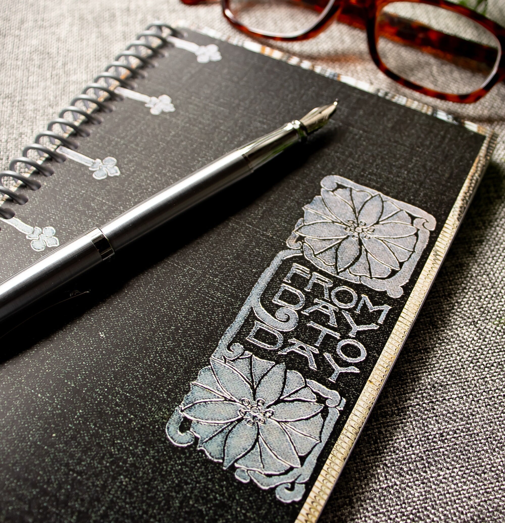 A travel-sized spiral notebook featuring a vintage book cover: FromDay To Day. Features a bold floral design. The colors are black, silver, gold and brown.