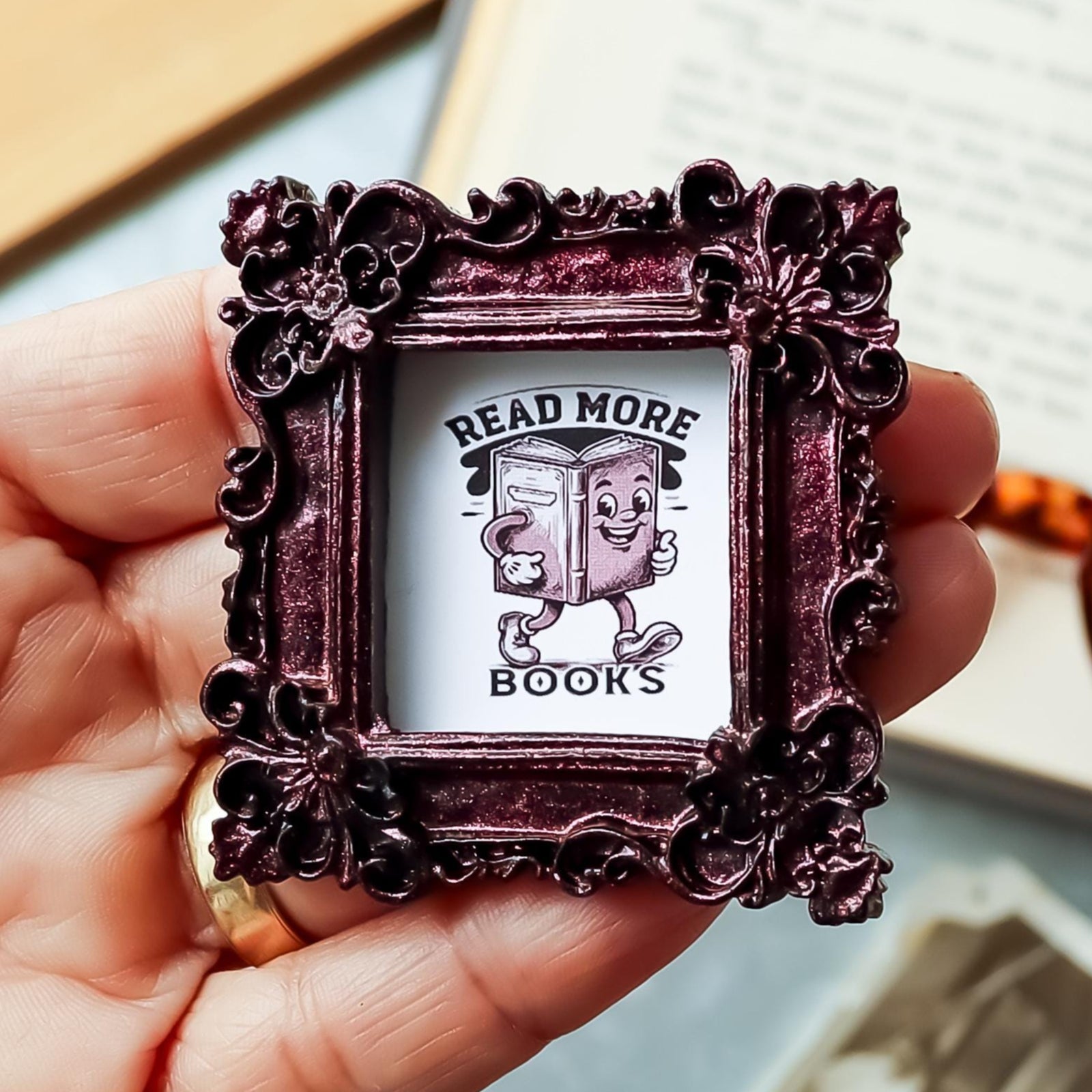 Hand holding &quot;Read More Books&quot; retro style refrigerator magnet. This is a small framed refrigerator magnet, featuring the phrase &quot;Read More Books&quot;, made by SunshineandRavioli. The resin frame is ornate vintage style in a metallic burgundy finish. A retro style illustration features a whimsical walking book giving a &quot;thumbs up&quot;.