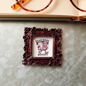 This is a small framed refrigerator magnet on a galvanized board, It features the phrase &quot;Read More Books&quot;, and is made by SunshineandRavioli. The resin frame is ornate vintage style with a metallic burgundy finish. It measures 2.25 x 2.25 inches.