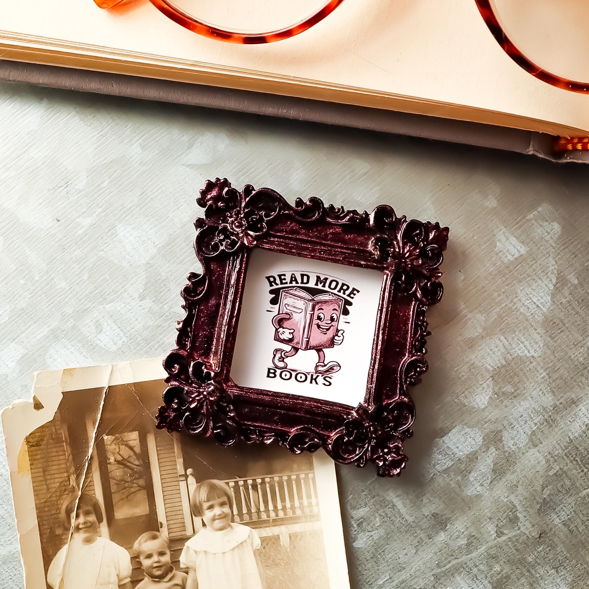 This is a small framed refrigerator magnet holding a photo on a galvanized board, It features the phrase &quot;Read More Books&quot;, and is made by SunshineandRavioli. The resin frame is ornate vintage style with a metallic burgundy finish. It measures 2.25 x 2.25 inches.