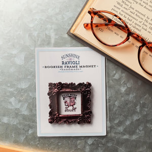 This shows packaging for a small framed refrigerator magnet that is made by SunshineandRavioli. The resin frame is ornate vintage style with a metallic burgundy finish. It measures 2.25 x 2.25 inches. The magnet is packaged in a small clear heat heat-sealed bag with a cardstock sleeve.